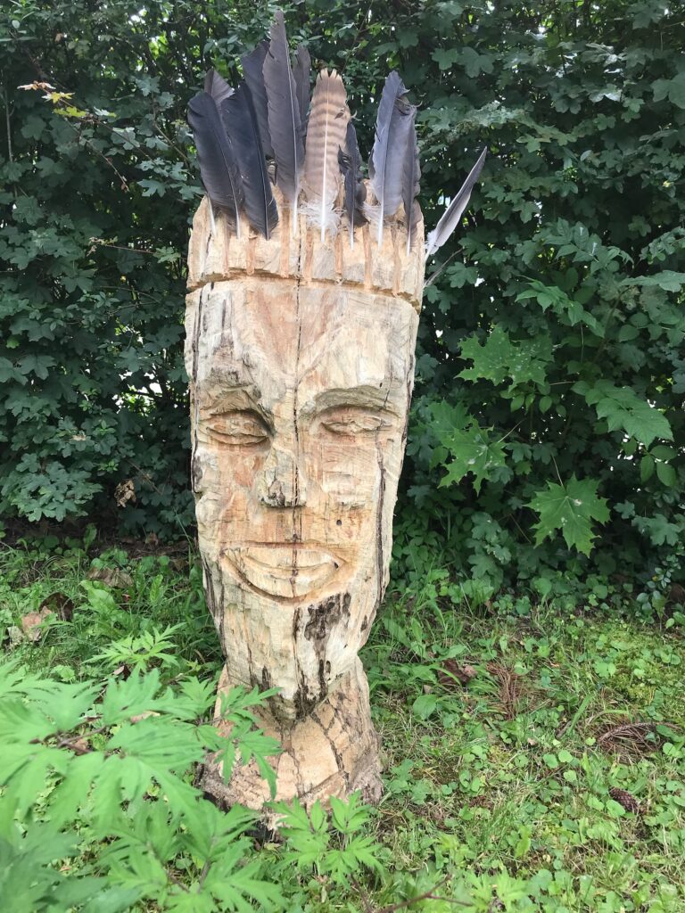 Holzskulptur eines Gesichts mit Federn um den Kopf.
